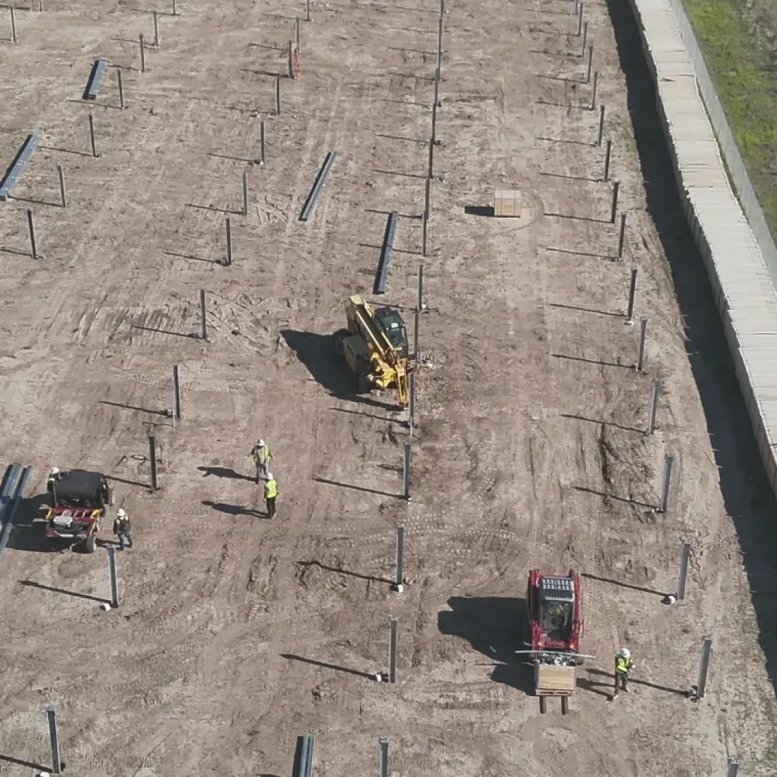 Aerial view of a construction site with rows of metal posts installed in the ground. Workers in high-visibility vests are operating machinery and handling materials. A dirt path runs alongside a green grassy area and a road is visible on the right.