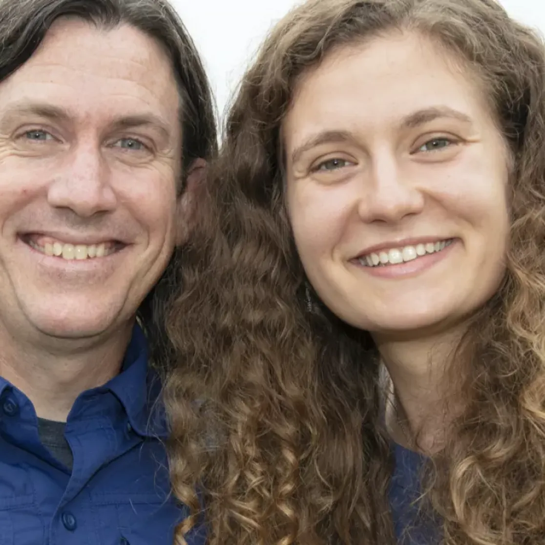 A man and woman smiling outdoors with a blurred background of trees and a building. The man has long hair and is wearing a blue shirt. The woman has curly hair and is also wearing a blue shirt.