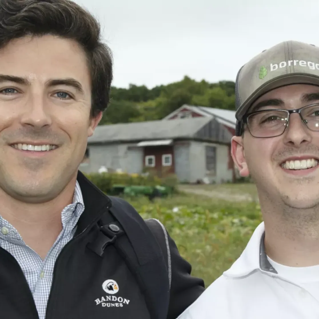 Two men smiling outdoors in front of a barn and trees. One wears a dark jacket with a logo, the other a cap and glasses.