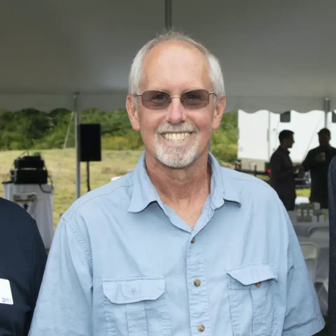 Three men standing under a tent at an outdoor event. The man on the left is wearing a navy polo shirt and glasses. The man in the middle is wearing a light blue shirt and sunglasses. The man on the right is wearing a dark suit jacket over a light blue shirt. They are all smiling, and there are people and tables in the background.