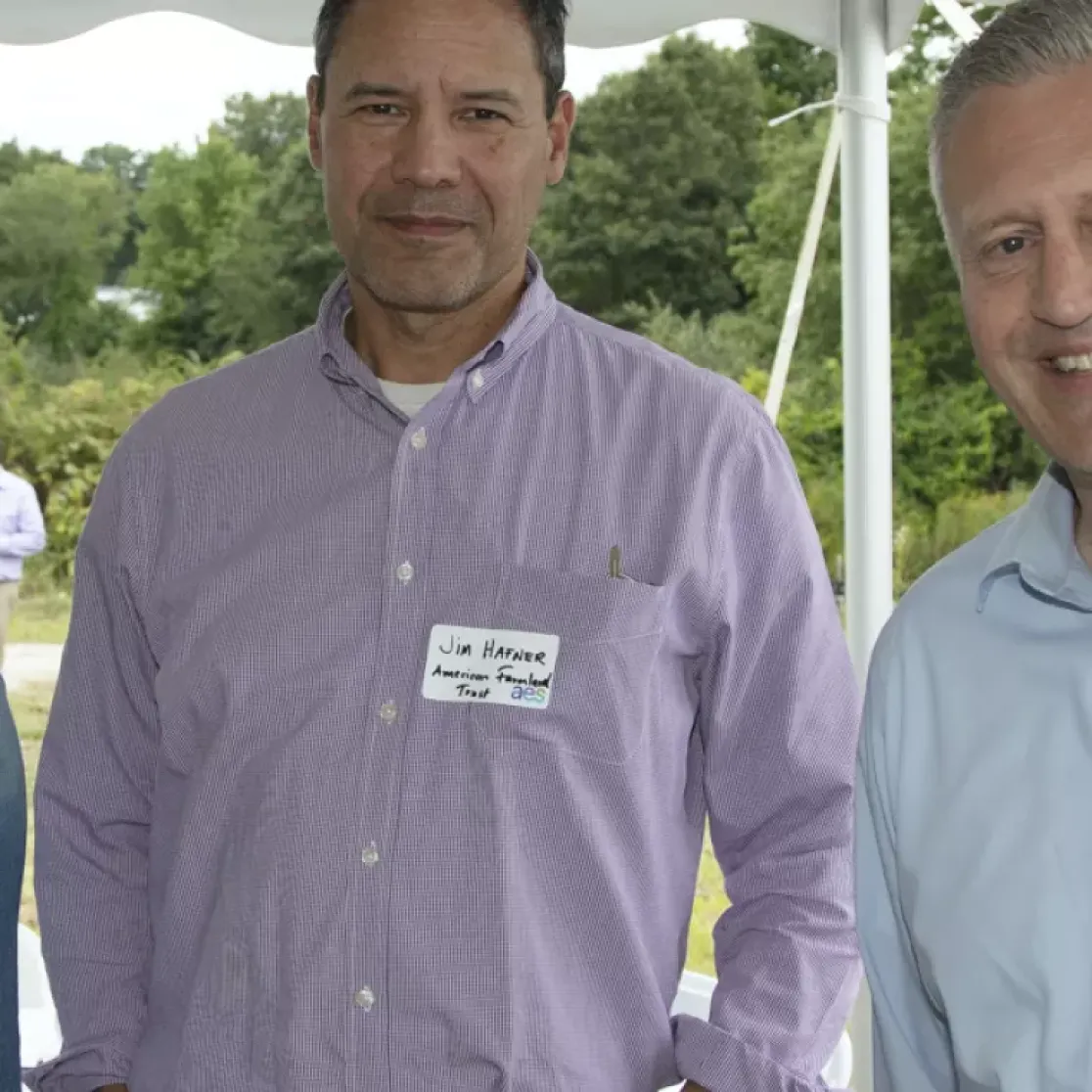 Three people standing under a tent at an outdoor event. The person on the left wears a denim shirt and sunglasses on their head. The middle person wears a purple shirt, and the person on the right wears a light blue shirt. A truck and greenery are visible in the background.