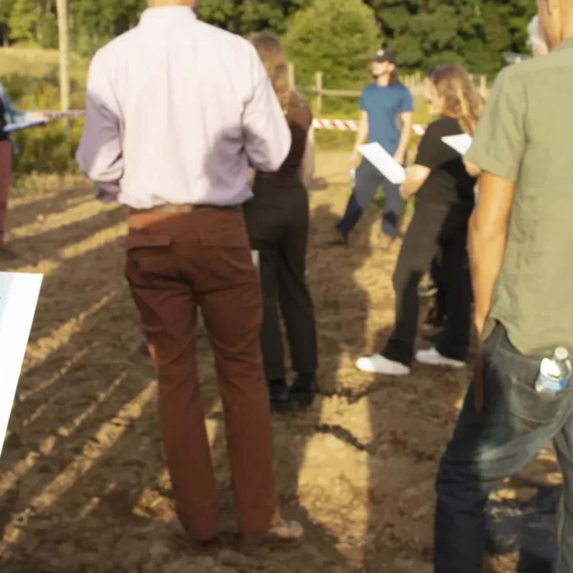 A group of people standing on a dirt path outdoors, holding papers and listening to a speaker. The scene is set near a field with greenery in the background. One person in the foreground holds a paper with a chart and text about impacts on agriculture.