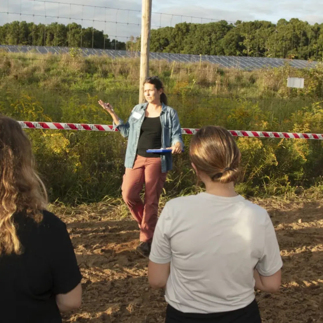 A group of people stands outdoors near a field with solar panels in the background. A woman is speaking to the group, gesturing with her hand. A red and white tape marked 'Danger Do Not Enter' separates the group from the field. Trees and greenery surround the area under a partly cloudy sky.