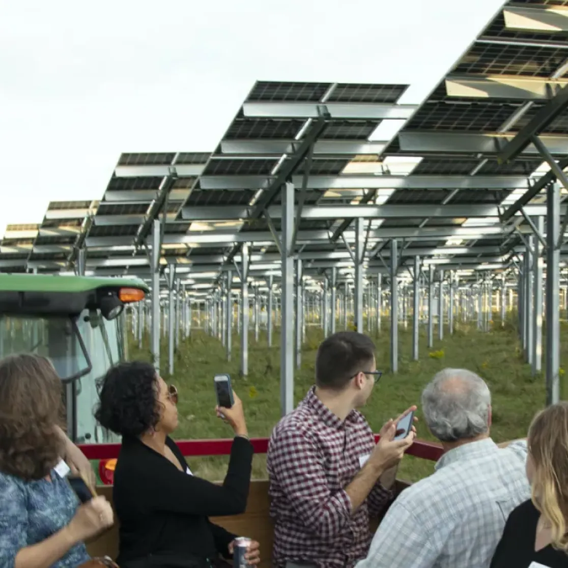 A group of people stand near a tractor, observing a field of elevated solar panels. The panels are arranged in rows, supported by metal structures. Trees are visible in the background under a clear sky.