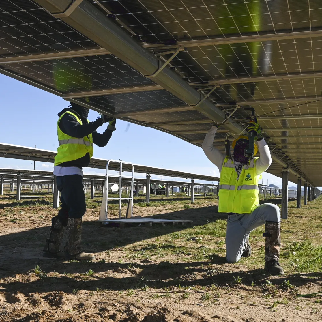 Three workers in safety vests and helmets install solar panels on a metal structure in an open field. The workers are using tools to secure the panels, with one worker kneeling and two standing. The sky is clear and blue, and rows of solar panels extend into the distance.