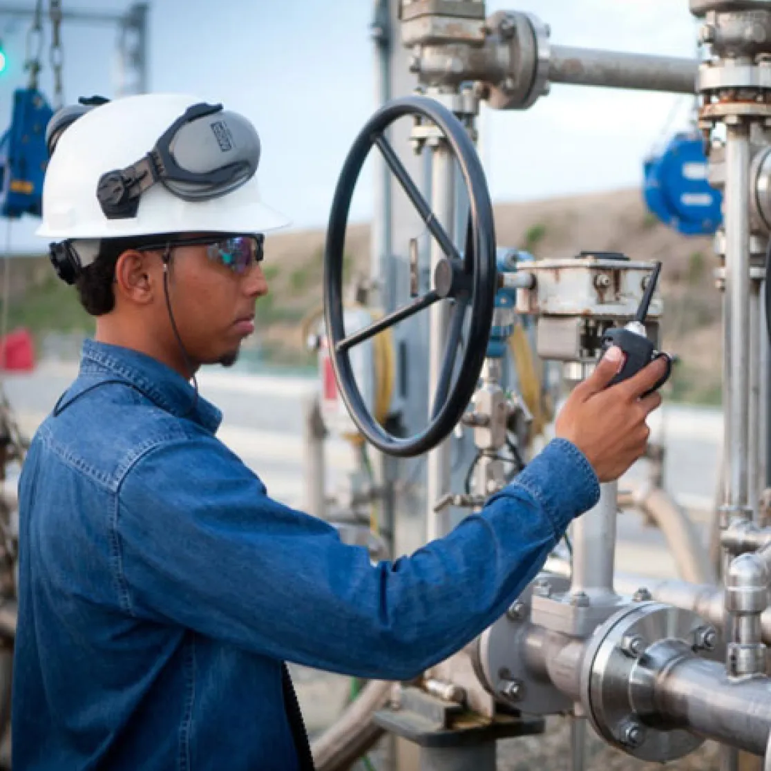 Worker in a hard hat and safety goggles operating a valve on industrial piping outdoors.