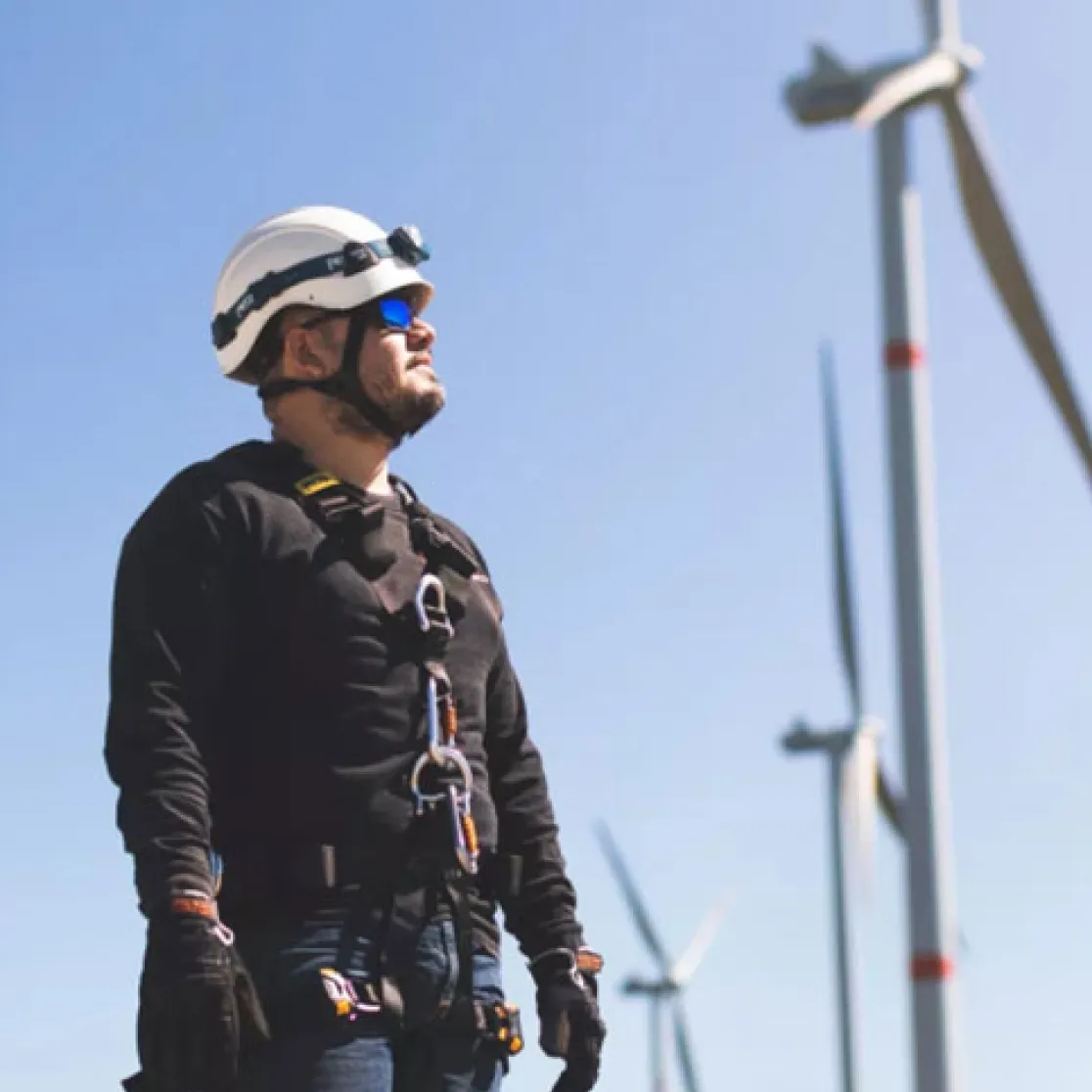 A worker wearing safety gear and a helmet stands in front of wind turbines on a clear day.