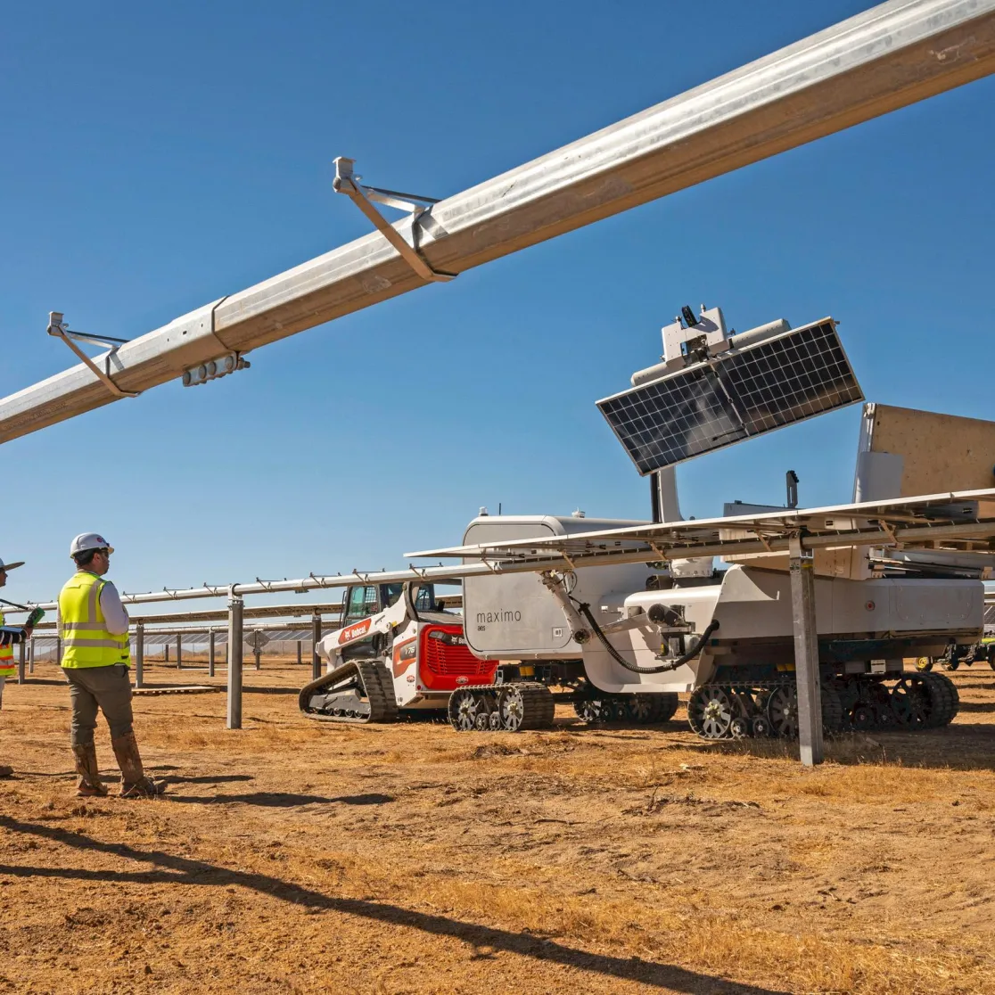 Three workers in safety gear stand near a solar panel installation site with a robotic vehicle on a sunny day.