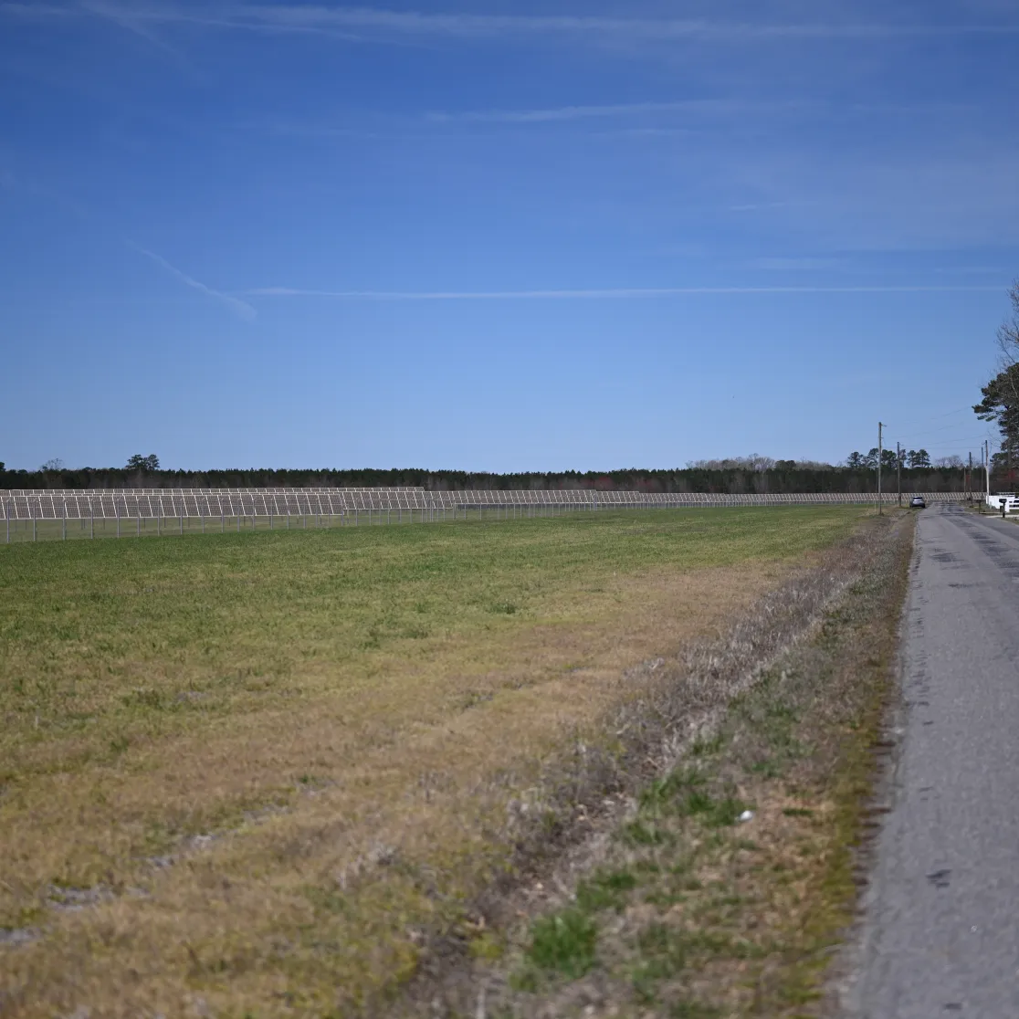 A rural landscape featuring a narrow paved road on the right and a large grassy field on the left. A row of solar panels is visible in the distance, parallel to the road. The sky is clear and blue, with some trees lining the horizon.