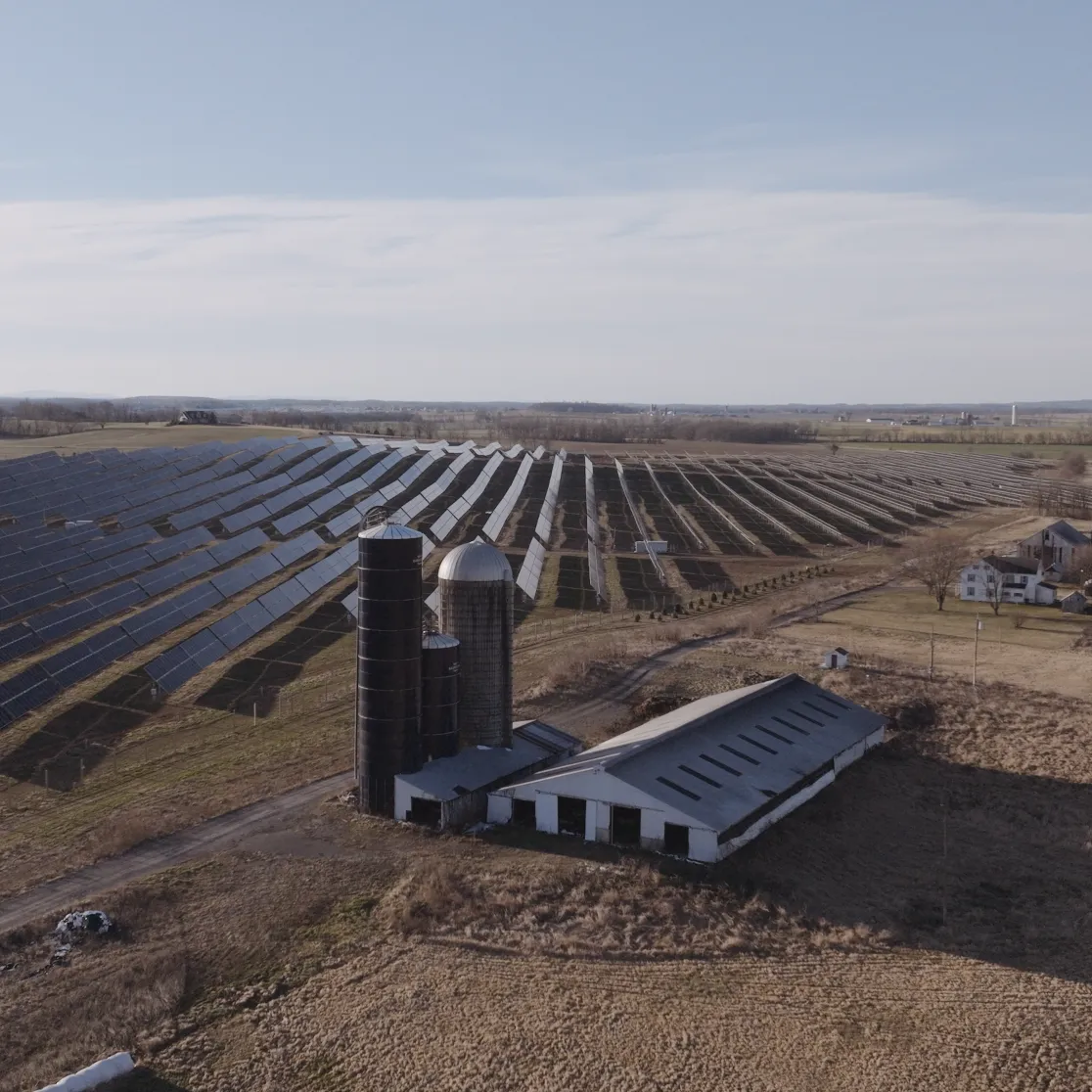 Aerial view of a large solar farm with rows of solar panels stretching across a vast field. In the foreground, there is a barn and silos. The landscape is rural with distant mountains under a clear sky.