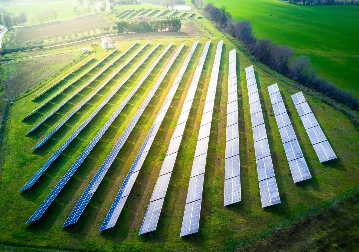 Aerial view of a solar farm with multiple rows of solar panels on a grassy field, surrounded by green countryside and trees.
