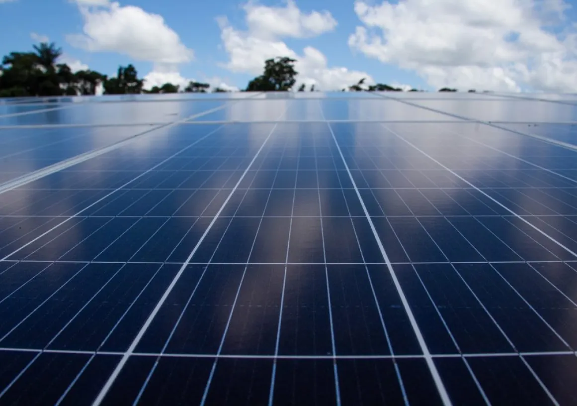 Close-up of a solar panel array under a blue sky with scattered clouds. The panels are aligned in rows, reflecting the sky and clouds above. Trees are visible in the distant background.