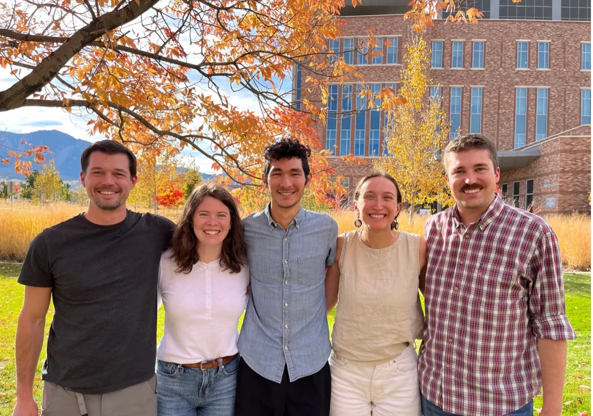 Five people standing together outdoors, smiling under a tree with autumn leaves. A brick building and mountains are visible in the background.