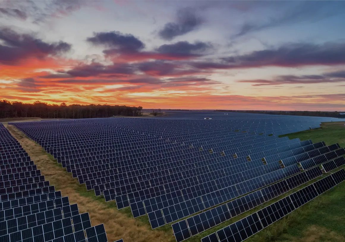 A vast field of solar panels under a vibrant sunset sky with red, orange, and purple hues.