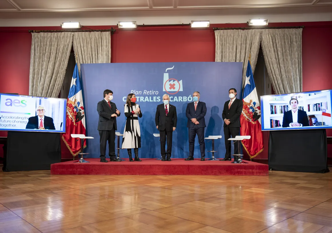 Five people wearing masks stand on a stage in front of a backdrop with the text 'Plan Retiro Centrales Carbón.' Two screens on either side display virtual participants. The setting is formal, with flags and curtains in the background.