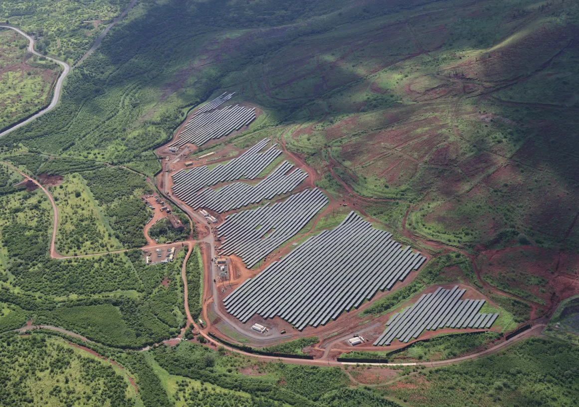 Aerial view of a large solar farm with rows of solar panels surrounded by lush green hills and a winding road.