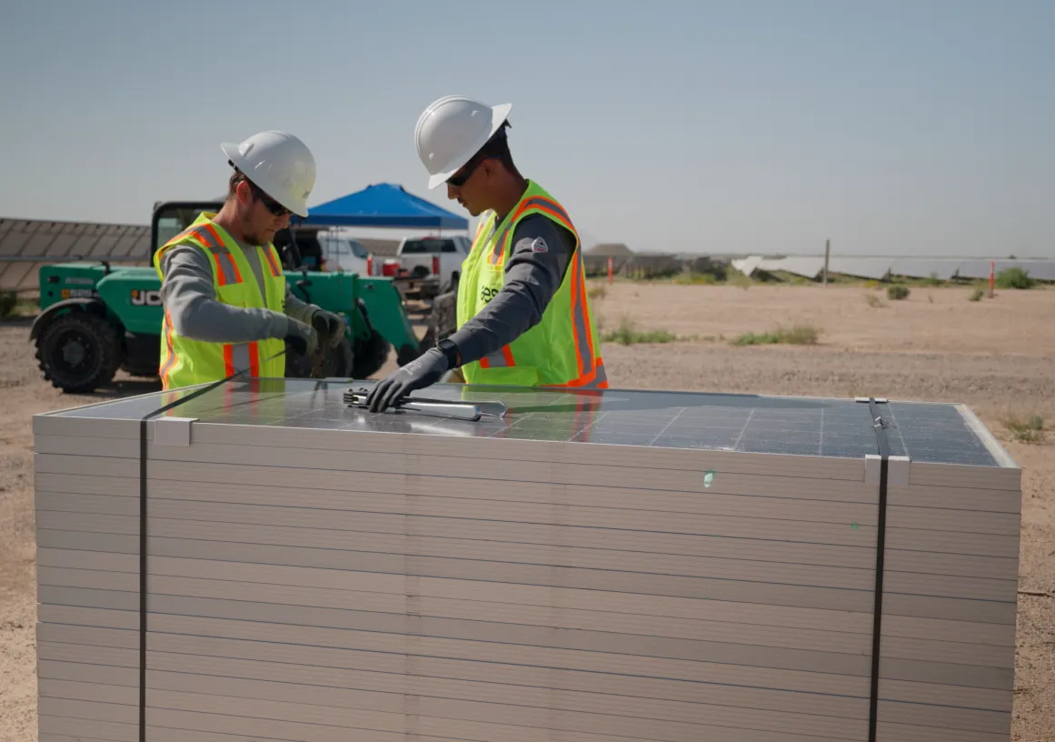 Two construction workers in safety vests and helmets inspect solar panels stacked on a pallet at a solar farm. A tractor and more solar panels are visible in the background under a clear sky.