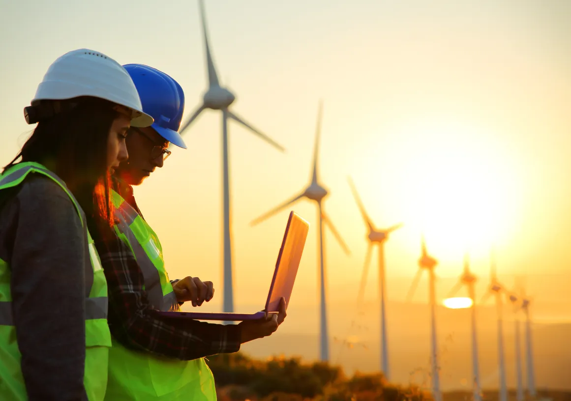 Two workers wearing hard hats and reflective vests stand in front of wind turbines at sunset. One holds a laptop, while the other looks on. The sky is a warm gradient of colors, and the wind turbines are silhouetted against the light.