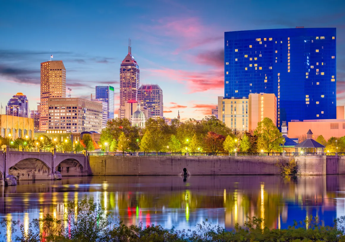 A city skyline at dusk with tall buildings, including a prominent blue glass structure, reflecting in a calm river. A bridge spans the river, and trees line the riverbank, illuminated by streetlights. The sky is a mix of blue and pink hues.