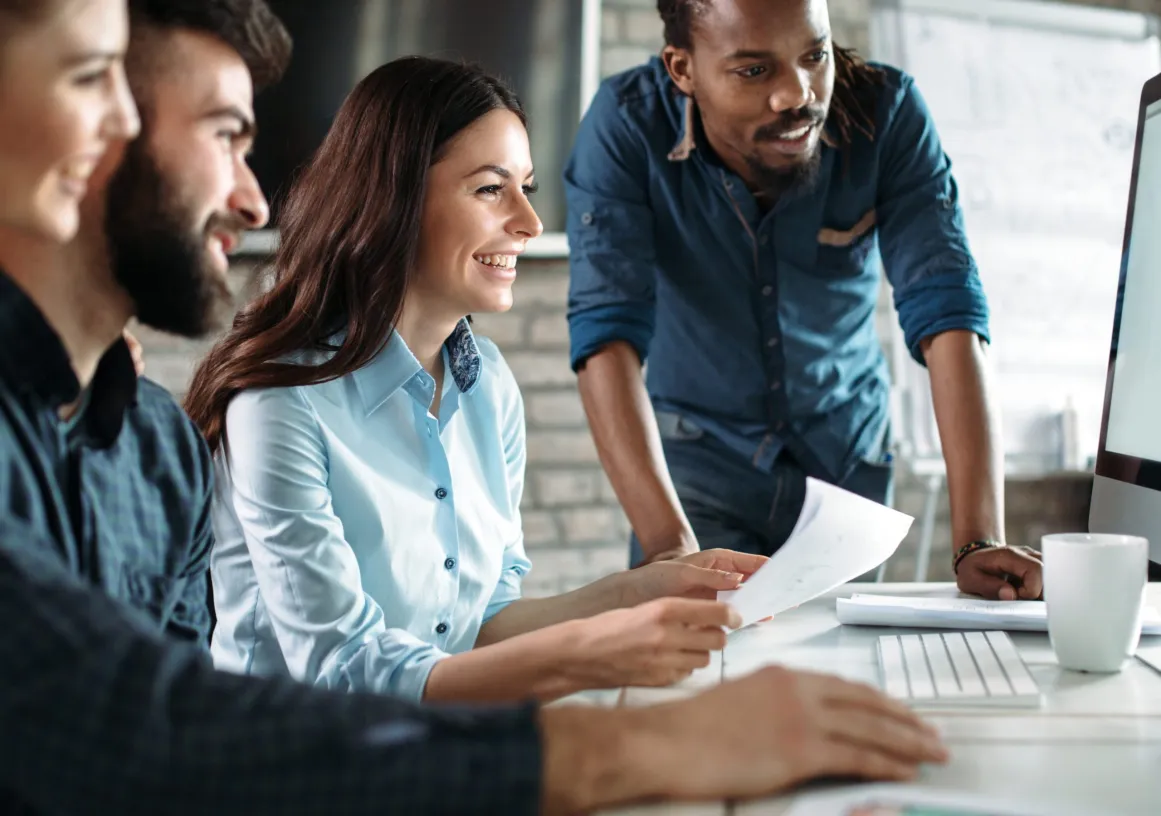 Four people in a modern office setting, sitting and standing around a computer screen, smiling and discussing. One person holds papers, and a whiteboard is visible in the background.