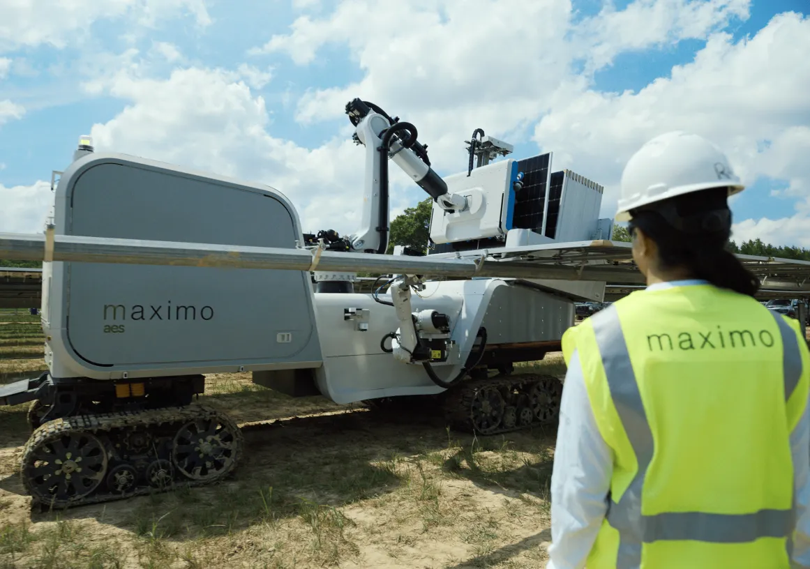 A person wearing a safety vest and hard hat observes a solar panel installation robot labeled 'maximo' on a sunny day.