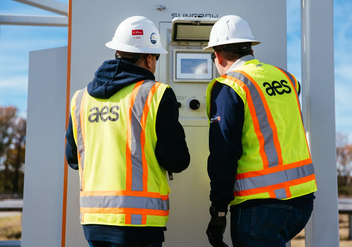 Two workers wearing white hard hats and yellow safety vests with 'aes' on the back are inspecting a control panel outdoors on a sunny day.