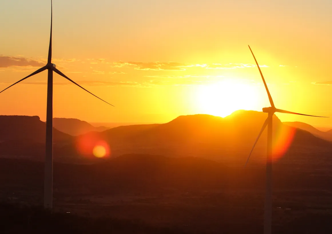 Two wind turbines silhouetted against a vibrant sunset with mountains in the background. The sky is filled with warm hues of orange and yellow, and the sun is partially visible behind the hills.