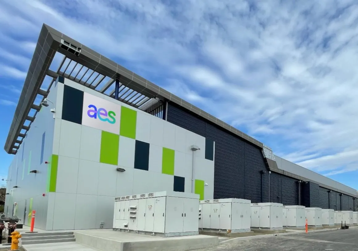 A modern industrial building with a sign displaying 'aes' on the facade. The building has a sleek design with green and dark panels. Several large electrical cabinets are positioned in front of it. The sky is partly cloudy.