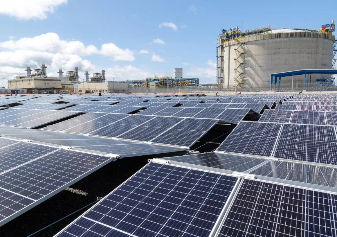A large field of solar panels under a blue sky, with an industrial facility and a large cylindrical structure in the background.