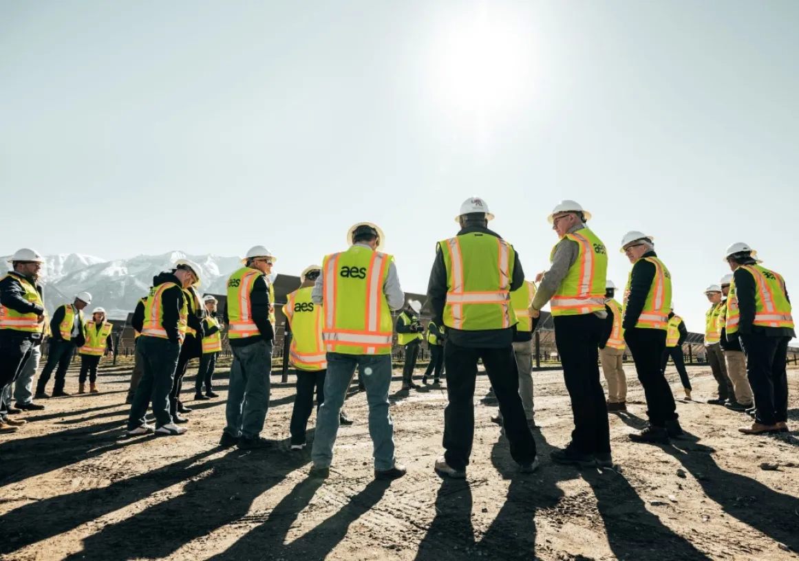 Group of construction workers in high-visibility vests and hard hats standing in a circle on a sunny day, with mountains in the background.