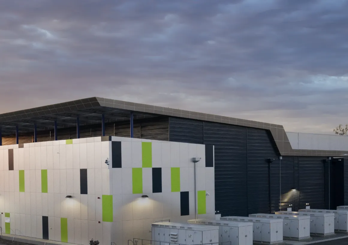 A modern industrial building with a colorful facade featuring green and brown panels, set against a cloudy sky at sunset. The building is surrounded by equipment and power lines.