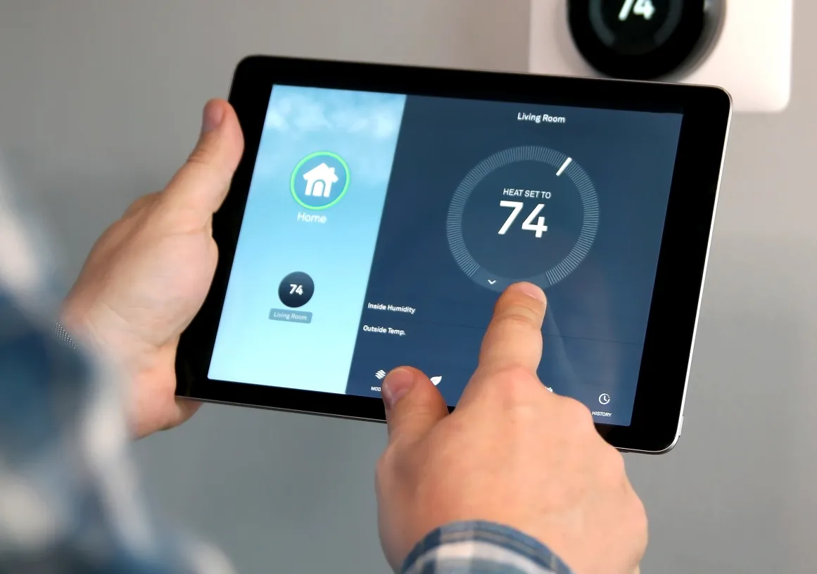 Person adjusting a smart thermostat using a tablet. The screen shows a temperature setting of 74 degrees for the living room. A wall-mounted thermostat displays the same temperature.