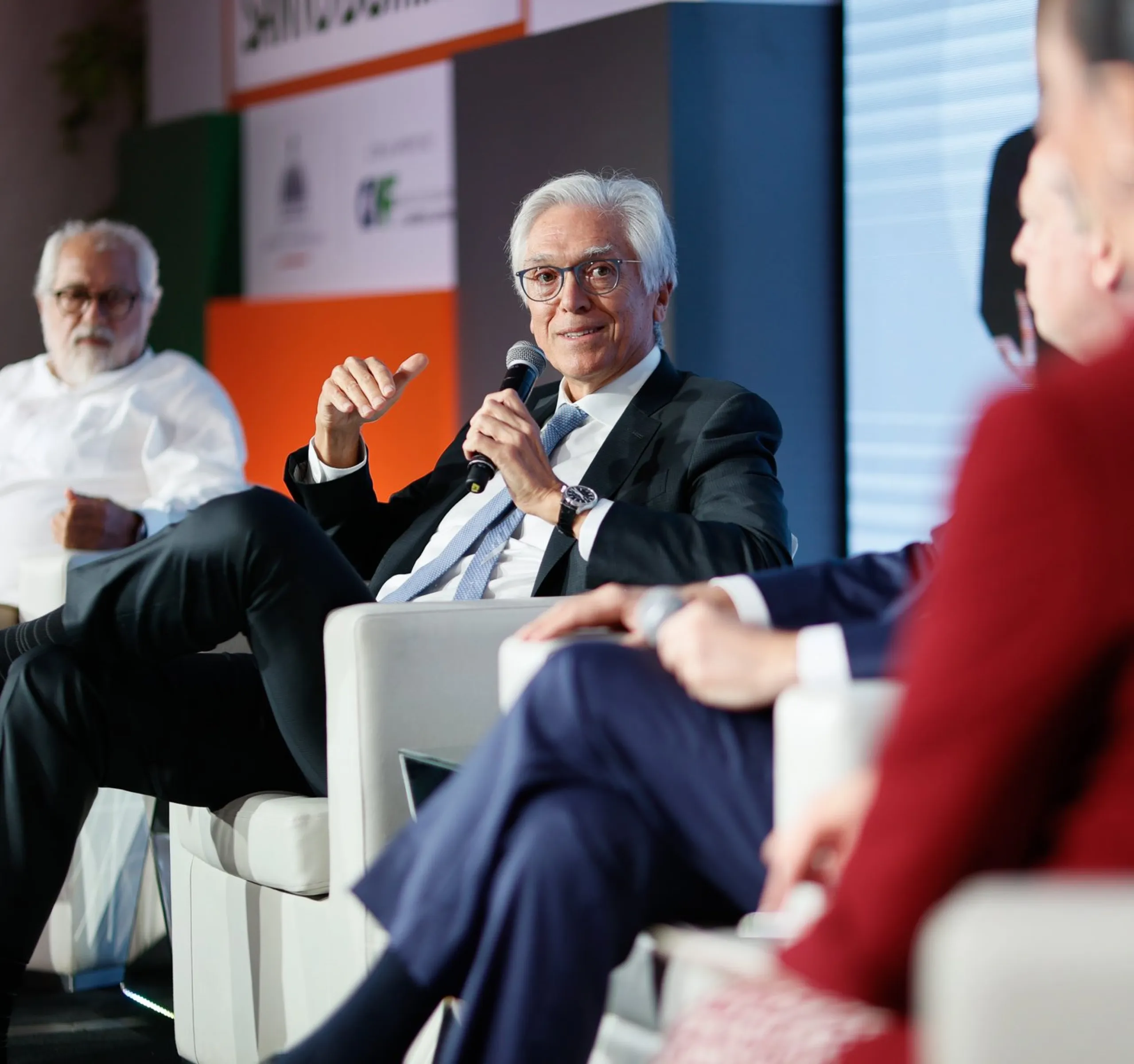 A panel discussion with four people seated in chairs. One person in a suit is speaking into a microphone. Another person in a white shirt is listening attentively. The background features a colorful banner.
