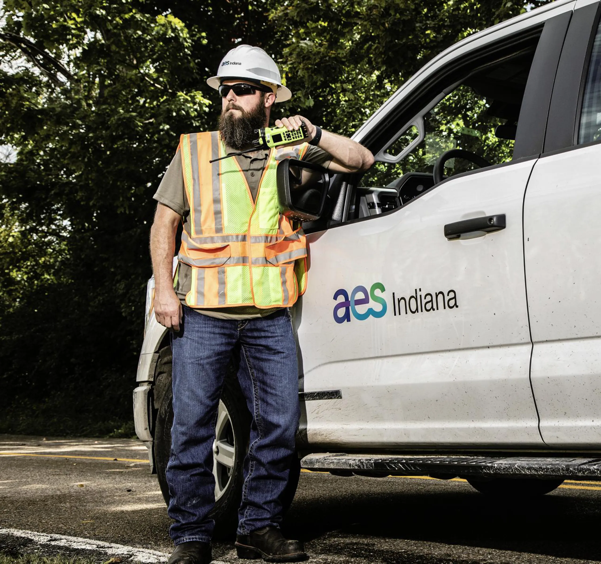 A person wearing a hard hat and reflective vest stands next to a white truck with 'AES Indiana' logo. They hold a device and lean on the truck's side mirror. Trees are visible in the background.