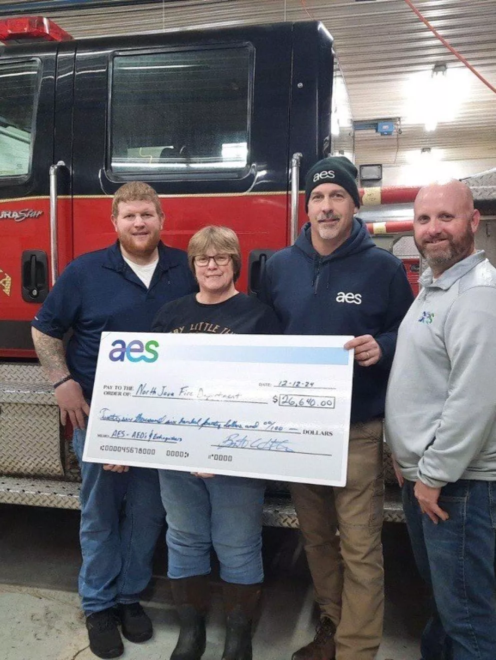 Four people stand in front of a North Java Fire Company fire truck holding a large check from AES for $26,640.00. The check is made out to the North Java Fire Department and dated 12-12-24.