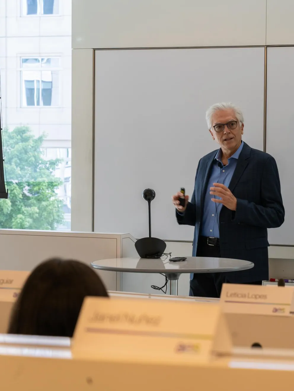 A man in a suit is giving a presentation in a conference room with a projector screen displaying an image and the AES logo. He stands near a round table with a microphone. Audience members are seated in front, with name tags visible. A window in the background shows a view of trees and a building.
