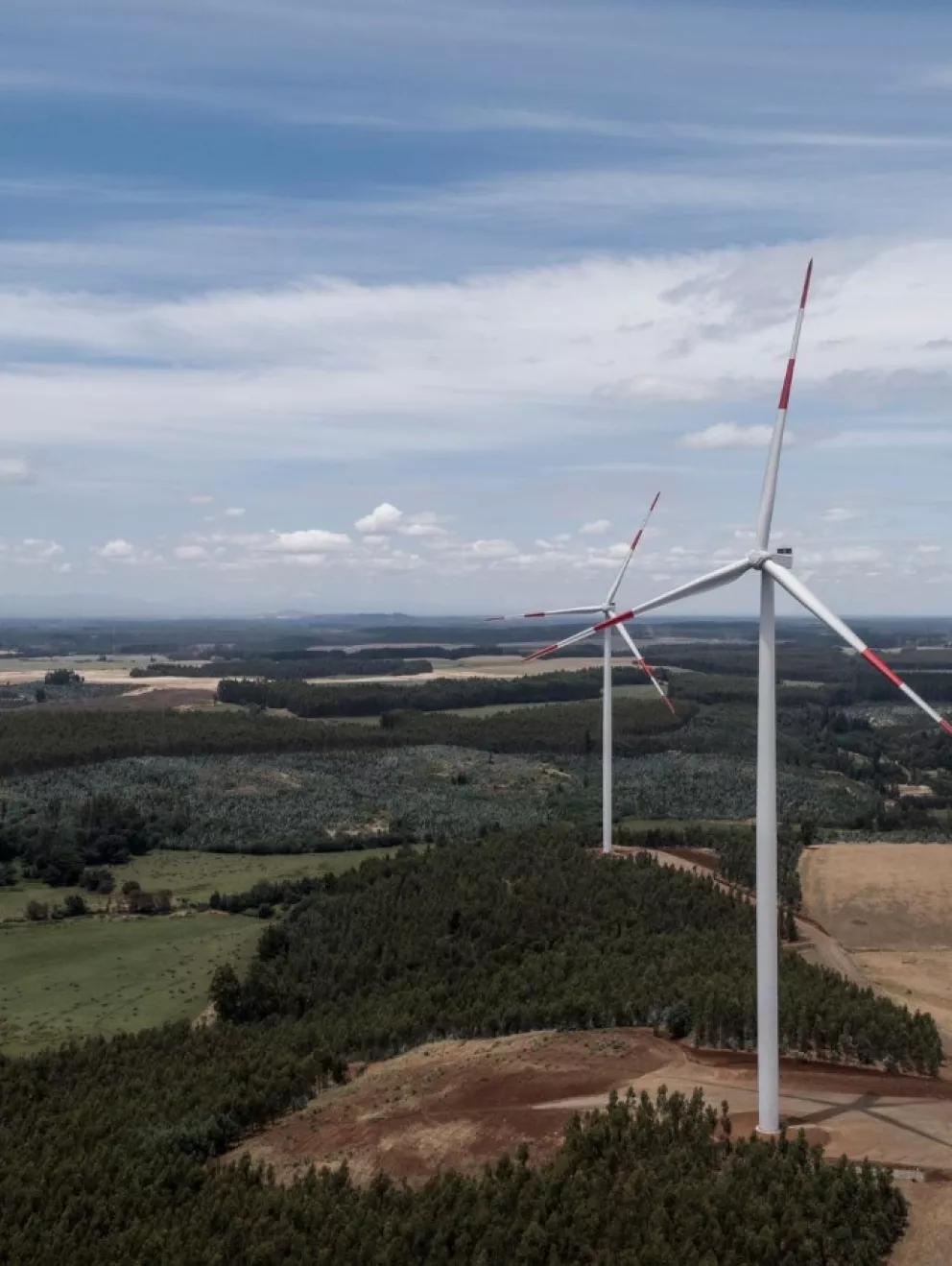 Aerial view of a landscape with two large wind turbines set amidst fields and scattered forests under a partly cloudy sky.