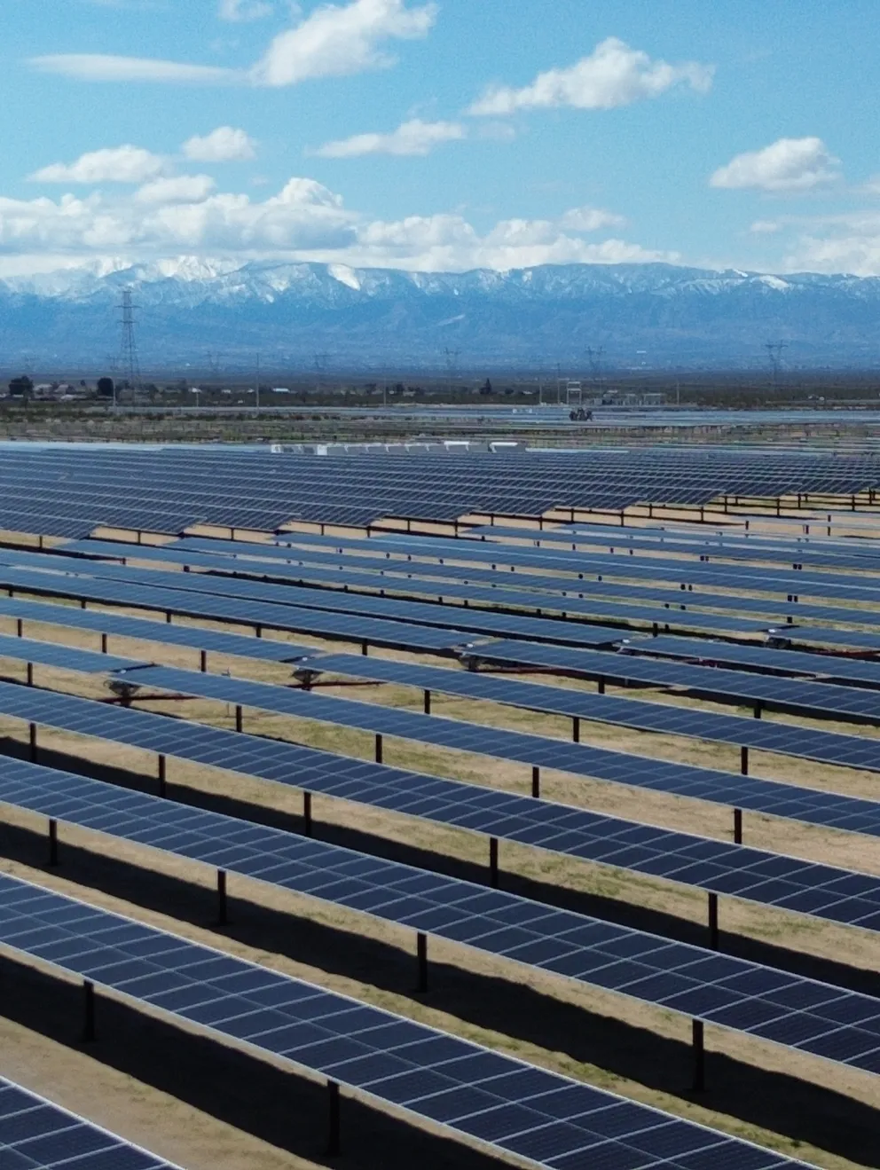 A vast solar farm with rows of solar panels stretching across a flat landscape. In the background, snow-capped mountains are visible under a partly cloudy sky.