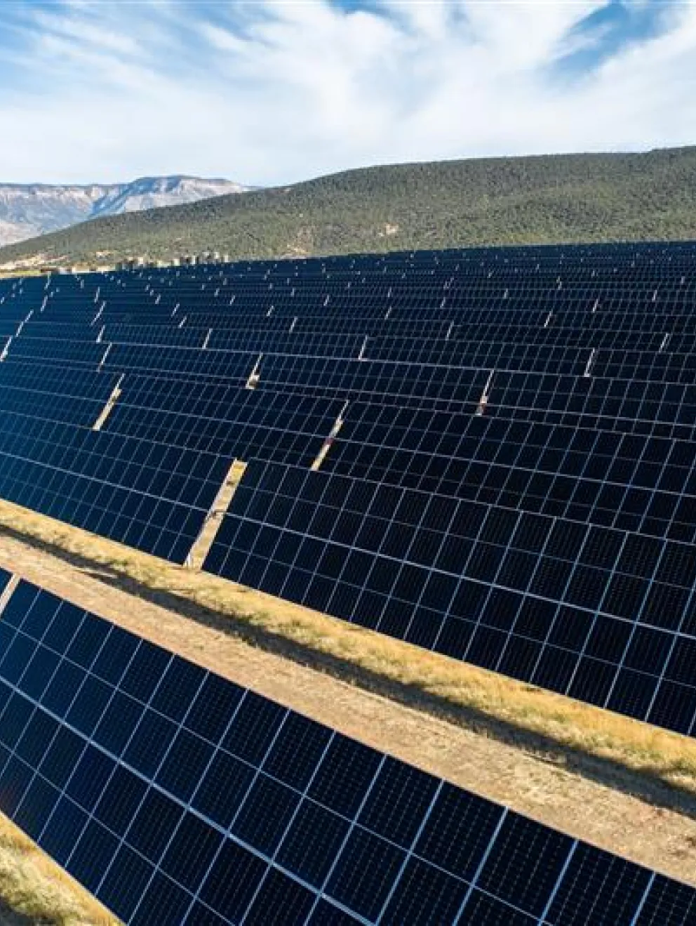 A large solar farm with rows of solar panels stretching across a dry landscape. The panels are aligned in neat rows, reflecting sunlight under a partly cloudy sky. In the background, there are hills and distant mountains.