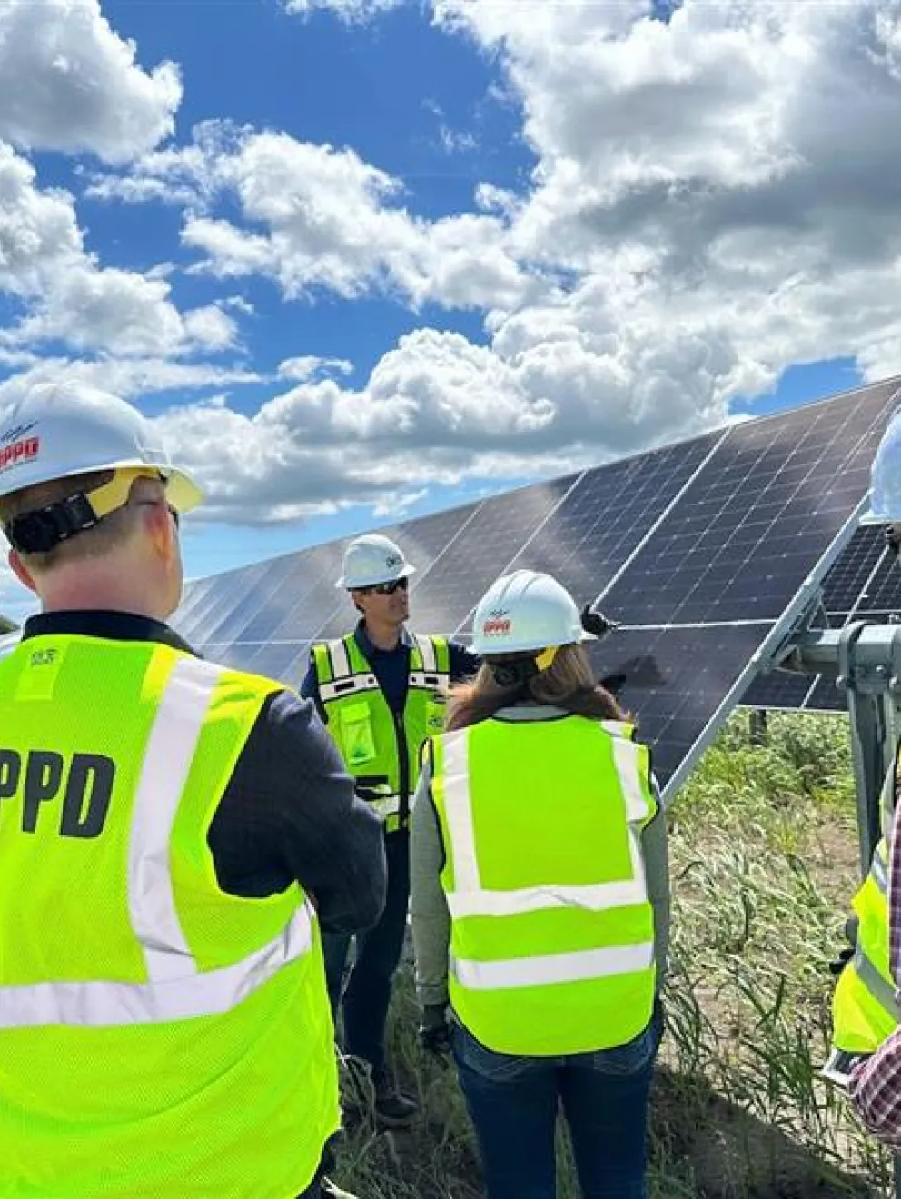 A group of people wearing hard hats and neon yellow safety vests stand in front of solar panels under a partly cloudy sky. They appear to be discussing or examining the solar equipment.