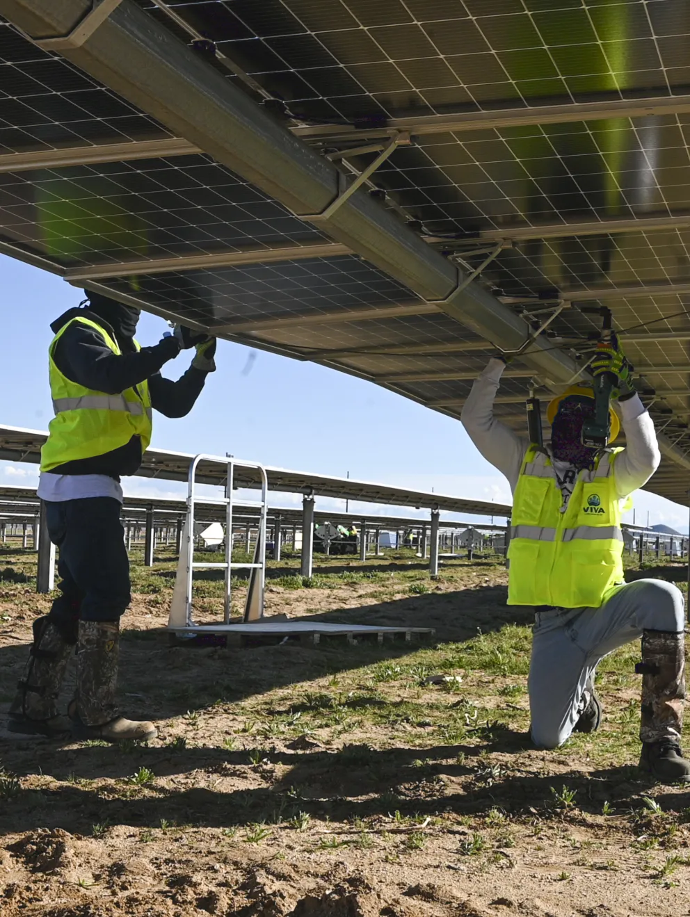 Three workers in safety gear and yellow vests install solar panels on a metal structure in a solar farm. One worker is kneeling, while the others stand on either side, all focusing on securing the panels.