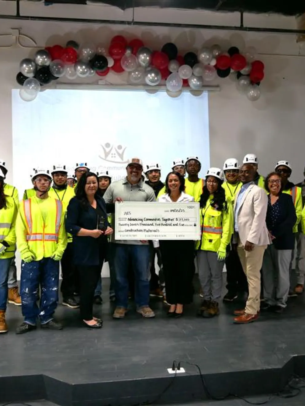 A group of people, including construction workers in safety gear and helmets, stand on a stage holding a large ceremonial check. The background features a screen and decorative balloons in red, black, and silver.
