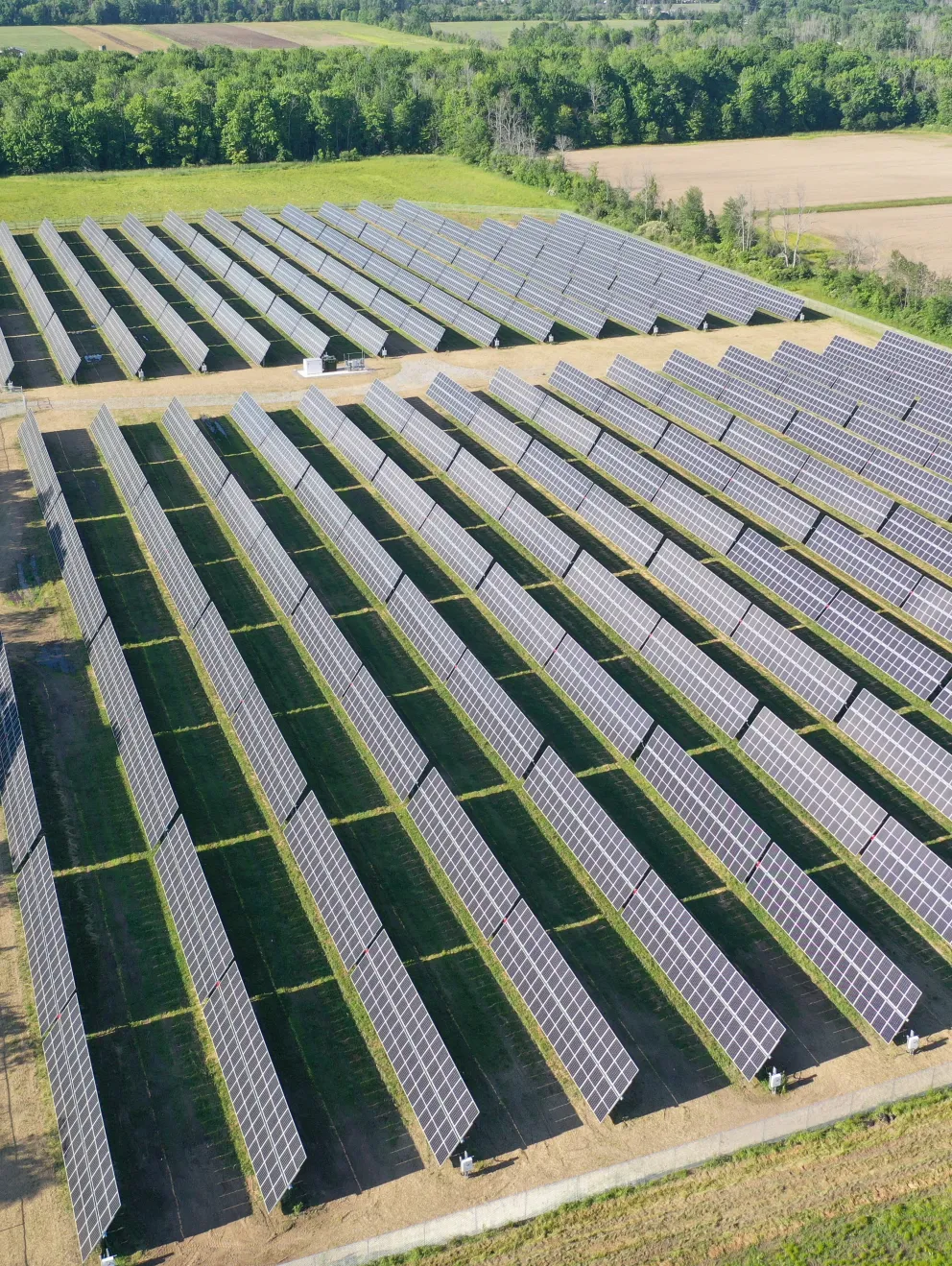 Aerial view of a large solar farm with rows of solar panels in a rural area surrounded by trees and fields.