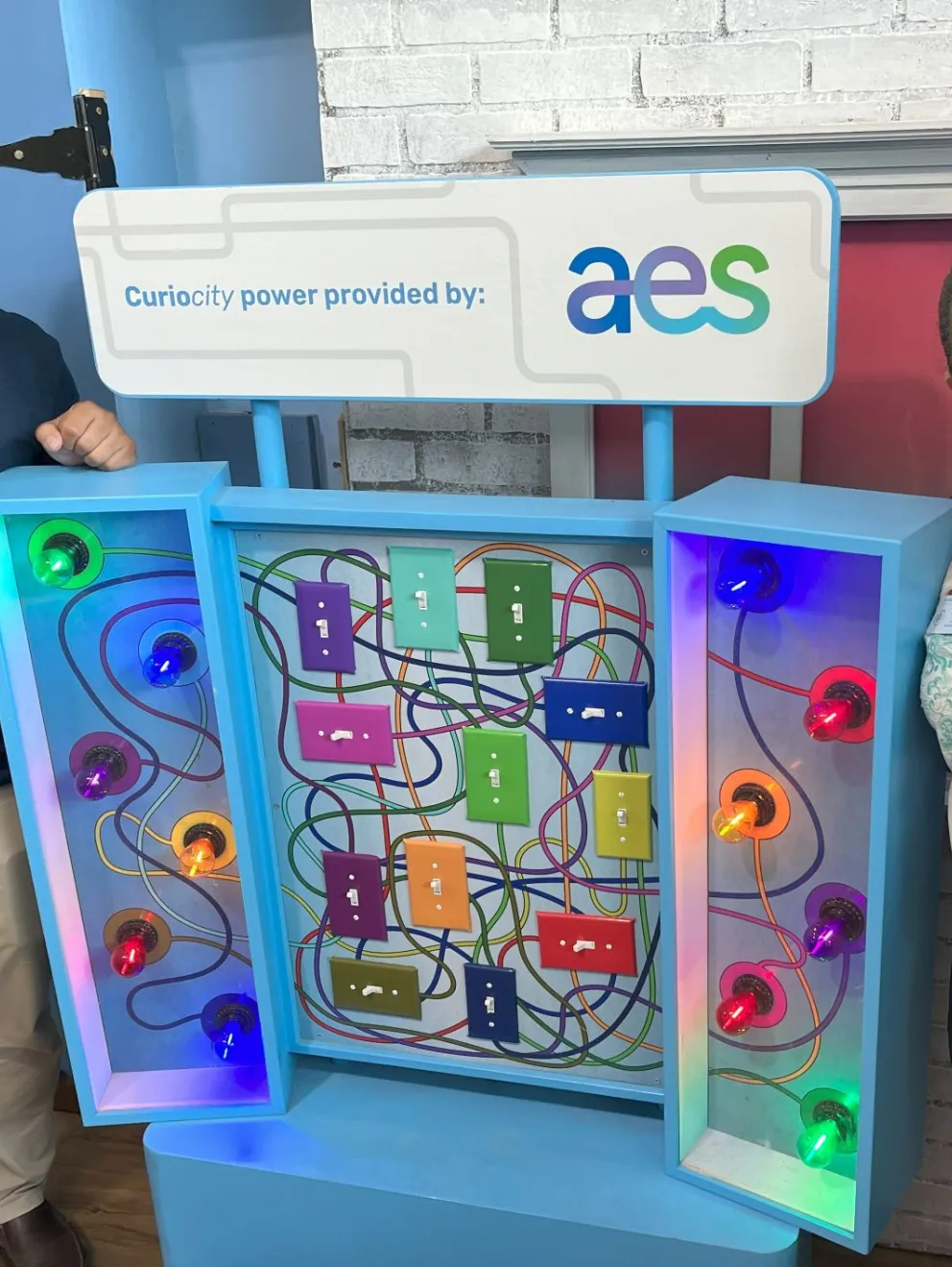 Two people stand beside a colorful interactive exhibit with various light bulbs and switches. The sign above reads 'Curiosity power provided by: aes'.