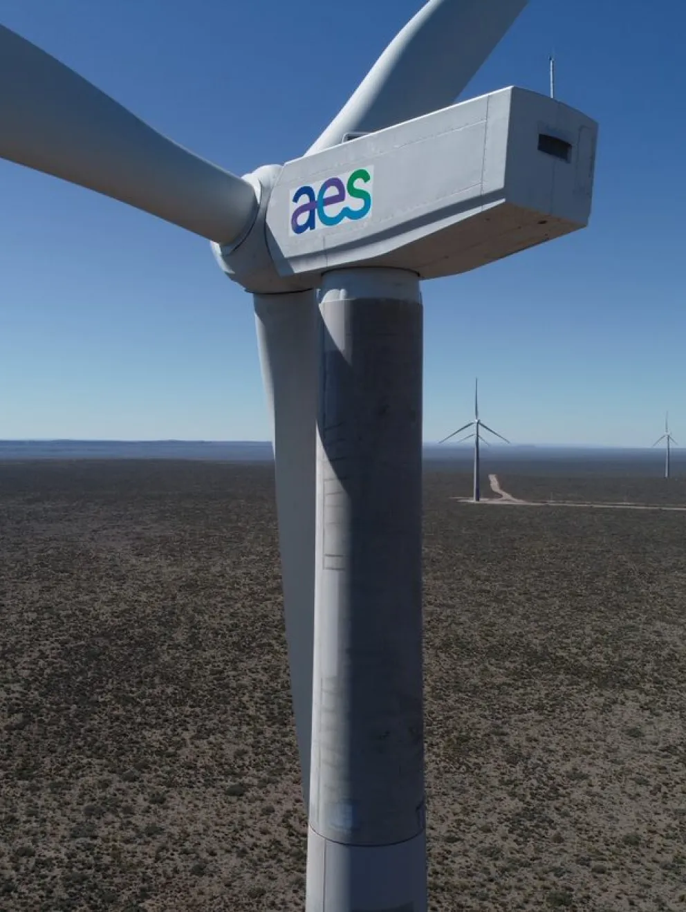 Close-up of a large wind turbine with the AES logo on its side, set in a vast open landscape with several other wind turbines in the distance under a clear blue sky.