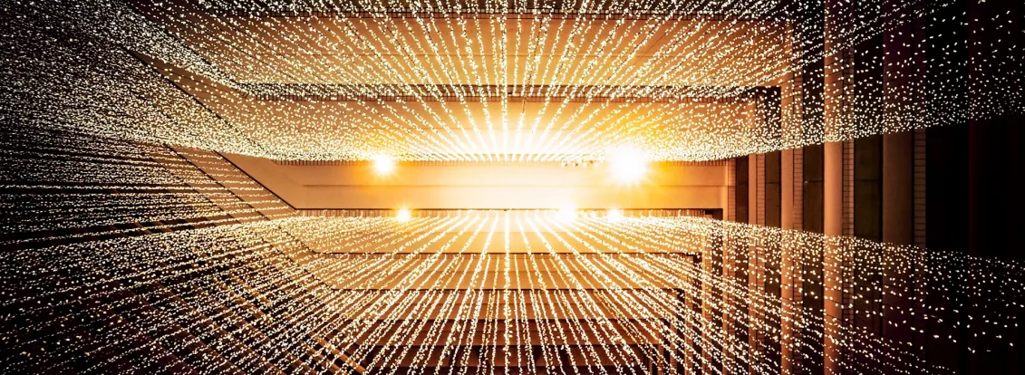 Rows of small white lights suspended in a geometric pattern inside a modern building, creating a radiant, starburst effect with warm lighting in the background.
