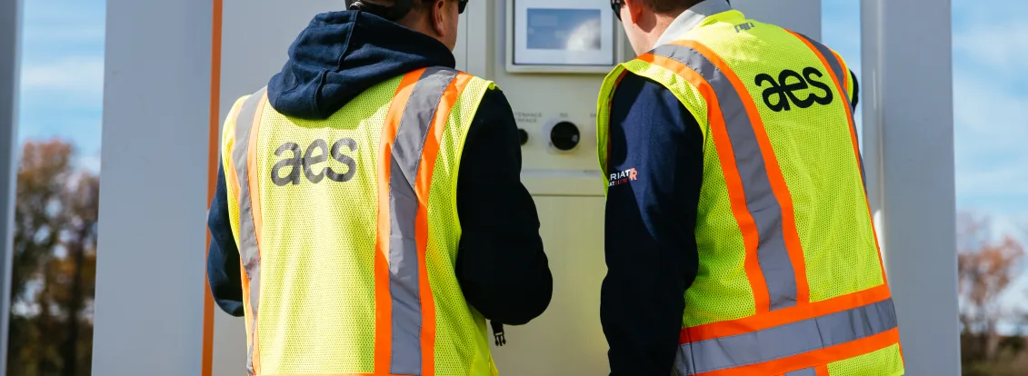 Two workers wearing white hard hats and yellow safety vests with 'aes' on the back are inspecting a control panel outdoors on a sunny day.