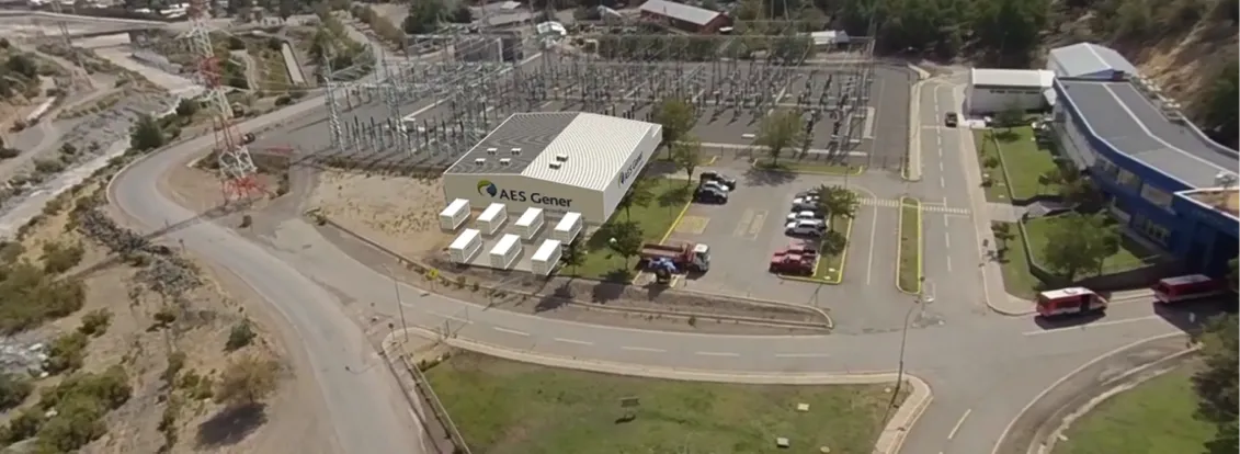 Aerial view of an industrial facility labeled 'AES Gener' surrounded by roads and greenery. The facility includes a main building, parking lot with several cars, and adjacent structures. Nearby, there is a substation with electrical equipment and power lines. The area is set in a mountainous landscape with trees and winding roads.