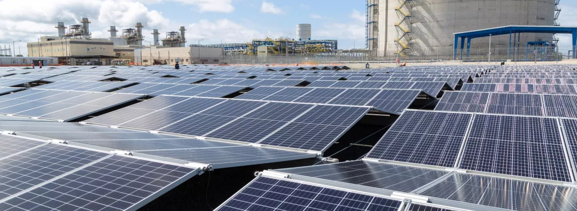 A large field of solar panels under a blue sky, with an industrial facility and a large cylindrical structure in the background.