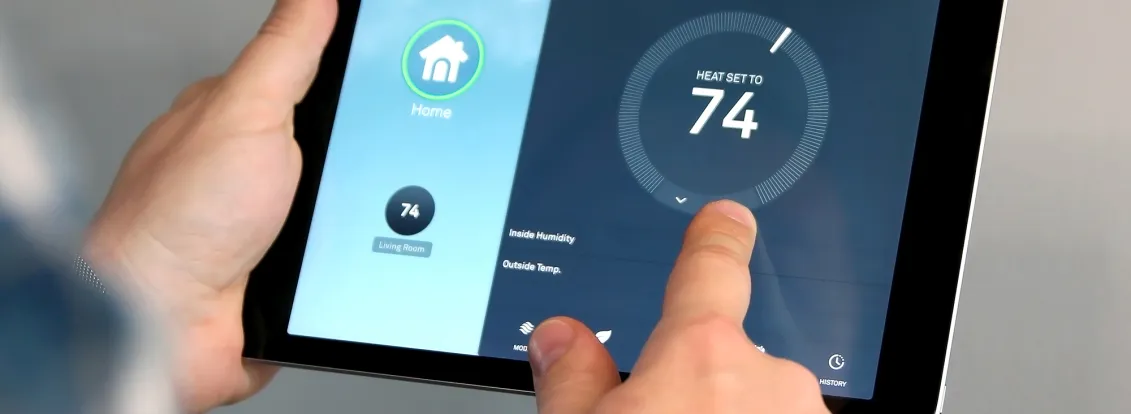 Person adjusting a smart thermostat using a tablet. The screen shows a temperature setting of 74 degrees for the living room. A wall-mounted thermostat displays the same temperature.